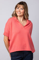 A woman modeling a coral pink short-sleeve knit polo shirt with a V-neck collar, paired with dark blue jeans, against a solid light gray studio background.