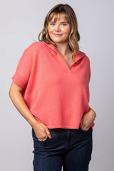 A woman wearing a coral pink short-sleeve polo shirt and dark blue jeans, standing against a light gray studio background.