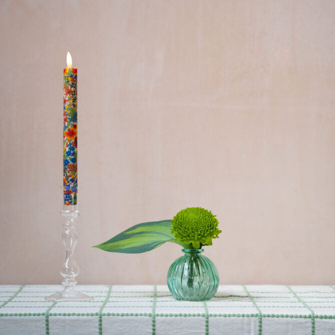 Decorative candle with floral design next to a small green vase with a leaf on a checkered tablecloth.